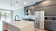 This thoughtfully designed one-bedroom, one-bathroom residence in Kannapolis, NC offers a sleek, modern aesthetic throughout its efficiently arranged single-floor layout. The open-concept kitchen and living area serves as the heart of the home, showcasing wide-plank hardwood flooring, soft neutral walls, and an abundance of natural light. The kitchen is a standout, featuring a large waterfall-edge island with an undermount sink and matte black fixtures, floor-to-ceiling dark wood cabinetry, a geometric tile backsplash, and a full suite of stainless steel appliances including a French door refrigerator and built-in microwave. Pendant lighting adds a refined touch overhead. The living room extends naturally from the kitchen and opens to a private outdoor balcony through expansive sliding glass doors, filling the space with light and outdoor views. The bedroom offers plush carpeting, a ceiling fan, and a window with natural light. The bathroom is finished with large-format taupe floor and wall tiles, a glass-enclosed shower, and matte black hardware accents that echo the home's contemporary design language. Clean lines and cohesive finishes create a polished, move-in-ready atmosphere throughout.