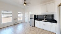 Located at 744 Beacon Avenue in Los Angeles, CA, this single-floor unit offers a compact yet functional layout with a clean, modern aesthetic. The space is finished with light hardwood-style flooring that runs throughout, contributing to a bright and cohesive feel.

The kitchen is a standout feature, equipped with white shaker-style cabinetry both above and below the counter, dark granite-look countertops, and a striking dark subway tile backsplash that adds contrast and visual interest. A stainless steel refrigerator and an integrated cooktop complement the space, along with a stainless steel sink fitted with a modern pull-down faucet.

The open living area is bathed in natural light courtesy of two large windows, which also provide views of the surrounding neighborhood. A wall-mounted heater unit is positioned between the windows for convenient climate control. A ceiling fan with a built-in light fixture is centrally mounted overhead, enhancing both air circulation and ambient lighting. The crisp white walls throughout lend the space a clean, airy atmosphere. A separate door leads to an additional room, providing functional separation within the layout. Overall, the unit presents a practical and neatly appointed space well-suited for urban living in the heart of Los Angeles.