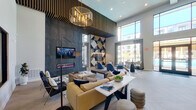 This contemporary single-floor community clubhouse is designed to impress, offering a dynamic blend of social, recreational, and relaxation spaces under soaring double-height ceilings throughout. The main lounge area is anchored by a dramatic dark stone feature wall housing a wall-mounted TV and a sleek linear electric fireplace, complemented by a bold geometric accent wall in navy, cream, and gold tones. Overhead, a striking slatted wood ceiling detail and a sculptural gold chandelier add architectural depth. Furnishings include plush caramel leather sofas, navy armchairs, and a substantial wood-block coffee table atop a layered area rug, creating an inviting and upscale atmosphere.

An adjacent social lounge opens to large floor-to-ceiling windows with pool views, bathing the space in natural light. Blue pod chairs and contemporary dining tables provide flexible seating arrangements. The herringbone light wood flooring transitions seamlessly into the game room, which features patterned geometric carpeting, a pool table, foosball table, a large-screen TV viewing nook with a plush navy sectional, and a prominent "GAME ON" wall sign with globe pendant lighting. Throughout, the space maintains a cohesive modern-urban aesthetic with curated artwork, high-end finishes, and abundant natural light.