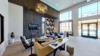 This contemporary single-floor community clubhouse is designed to impress, offering a dynamic blend of social, recreational, and relaxation spaces under soaring double-height ceilings throughout. The main lounge area is anchored by a dramatic dark stone feature wall housing a wall-mounted TV and a sleek linear electric fireplace, complemented by a bold geometric accent wall in navy, cream, and gold tones. Overhead, a striking slatted wood ceiling detail and a sculptural gold chandelier add architectural depth. Furnishings include plush caramel leather sofas, navy armchairs, and a substantial wood-block coffee table atop a layered area rug, creating an inviting and upscale atmosphere.

An adjacent social lounge opens to large floor-to-ceiling windows with pool views, bathing the space in natural light. Blue pod chairs and contemporary dining tables provide flexible seating arrangements. The herringbone light wood flooring transitions seamlessly into the game room, which features patterned geometric carpeting, a pool table, foosball table, a large-screen TV viewing nook with a plush navy sectional, and a prominent "GAME ON" wall sign with globe pendant lighting. Throughout, the space maintains a cohesive modern-urban aesthetic with curated artwork, high-end finishes, and abundant natural light.