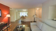 This single-floor residence offers a welcoming and functional layout characterized by an open-concept design that seamlessly connects the living, dining, and kitchen areas. Rich, dark hardwood flooring flows throughout the main living space, adding warmth and elegance to the interior. The living area is bright and inviting, anchored by a white sofa, a stylish floor lamp, and a coffee table, all set atop a plush area rug. A white spindle railing detail adds a charming architectural accent to the space. The kitchen features a raised breakfast bar with seating for two bar stools, serving as a casual dining spot as well as a natural divider between the cooking and living areas. A window above the breakfast bar allows natural light to pour in, creating a bright and airy atmosphere. Soft, neutral tones on the walls complement the warm flooring throughout, lending the space a cohesive and clean aesthetic. Recessed and flush-mount lighting provides consistent illumination throughout. A hallway visible from the main living area leads to additional rooms, suggesting a practical and well-organized floor plan. Overall, this residence balances comfort and style in a thoughtfully designed single-level layout.