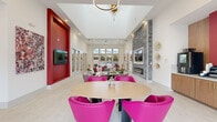 Located in Orlando, FL, this stylish single-floor community clubhouse delivers a generously sized, thoughtfully designed gathering space that blends bold aesthetics with practical amenities. The interior showcases soaring ceilings, large windows, and glass doors that flood the space with natural light while framing views of the outdoor pool area beyond.

The main lounge area features a dramatic stone accent wall with a linear fireplace, a wall-mounted TV set against a vivid crimson panel, and a mix of plush white sectionals and vibrant fuchsia accent chairs arranged around a round dining table. A striking circular gold chandelier anchors the center of this zone.

A dedicated media lounge offers expansive modular seating in deep red and cream tones, a ceiling-mounted projector aimed at a large projection screen, and a bold abstract mural that commands an entire wall. Pendant lighting and framed artwork add artistic depth throughout.

The wet bar area features warm wood cabinetry, a white quartz island with bar seating, pendant lights, and a mounted TV — ideal for social gatherings. A separate refreshment station includes a professional coffee machine, beverage refrigerator, and gold-accented cabinetry set against a crimson accent wall.

Light hardwood-style flooring, textured area rugs, and curated gold wall sculptures complete the sophisticated, resort-inspired atmosphere of this vibrant community space.