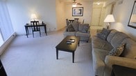 This single-story space showcases an open-concept layout that seamlessly connects the living and dining areas, creating a cohesive and practical living environment. The living area is anchored by plush wall-to-wall carpeting in a neutral tone, providing a comfortable and inviting atmosphere. A ceiling fan with integrated lighting serves as a functional central fixture, ensuring year-round comfort and adequate illumination throughout the space.

The seating arrangement features a generously sized sectional and loveseat in a warm gray upholstery, accented with decorative throw pillows. A dark-finished rectangular coffee table serves as the focal point of the seating area, complemented by a matching console table along the wall that adds both style and additional surface space. Framed artwork above the loveseat adds a decorative touch to the neutral walls.

The dining area, positioned adjacent to the living space, accommodates a round table with several chairs, making it well-suited for everyday dining. Light tile flooring distinguishes the dining zone from the carpeted living area. A doorway visible at the rear of the space leads to additional rooms, rounding out this functional and well-arranged single-floor layout.