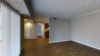 This single-floor residence offers a clean and functional layout designed for comfortable everyday living. The main living area is finished with rich, dark hardwood-style flooring that runs throughout, providing a warm and cohesive foundation across the space. Neutral gray walls create a versatile backdrop, while white crown molding adds a subtle decorative touch along the ceiling line. The textured ceiling contributes a classic residential character to the interior.

The open layout allows for a natural flow from the living area toward the kitchen and hallway beyond. The kitchen is visible from the main living space and features warm wood cabinetry, a dark countertop, and space for appliances, offering a practical and well-appointed cooking area. A hallway extends from the main living space, leading to additional rooms and providing clear connectivity throughout the home.

Recessed lighting in the hallway and an overhead vent in the living area speak to the functional infrastructure of the space. The overall ambiance is understated and move-in ready, with a neutral color palette and consistent flooring that lend the home a tidy, well-maintained feel suited for a variety of interior styles and furnishing choices.