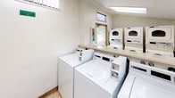 This shared laundry facility is a single-story, self-contained space designed for practical, communal use. The room is equipped with multiple top-loading washing machines, including Maytag models, as well as several stackable dryer units positioned along the walls, providing ample laundry capacity. A continuous laminate folding counter runs along one wall, offering a convenient surface for sorting and folding laundry. The flooring consists of neutral tile throughout, contributing to a clean, low-maintenance environment. Natural light fills the space through a series of wide horizontal windows positioned along the upper portion of the walls, offering views of the surrounding landscaped grounds, including greenery and adjacent brick buildings. Fluorescent overhead lighting supplements the natural light, ensuring the room remains well-lit at all times. A ventilated entry door with a window panel provides access to the exterior. A fire extinguisher cabinet is mounted near the entry for safety. The overall layout is efficient and organized, with white walls and light-colored surfaces maintaining a bright, utilitarian ambiance suitable for shared residential use.