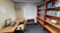 This single-floor office space is thoughtfully arranged to support a productive work environment. The room features an L-shaped wooden desk that provides generous workspace, paired with a comfortable tan leather executive chair. A wall-mounted air conditioning unit ensures climate control year-round, while a window dressed with vertical blinds allows for adjustable natural light throughout the day.

A prominent wooden bookshelf unit lines one wall, offering multiple shelves filled with binders, books, and reference materials, making it ideal for organized storage and easy access to documents. The shelving extends to the ceiling, maximizing vertical storage capacity. Additional lower shelving provides further space for materials and supplies.

The flooring transitions between carpet and a hard surface, defining the different functional zones within the room. Crown molding along the ceiling adds a subtle finishing detail to the otherwise utilitarian space. A small plant atop the shelving introduces a touch of greenery to the interior. Overall, the office conveys a practical and organized atmosphere, well-suited for focused work, research, or administrative tasks.
