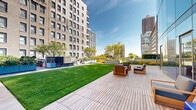 This expansive rooftop terrace offers a remarkable urban retreat, seamlessly blending lush landscaping with well-appointed outdoor living areas. The space is anchored by a generously sized manicured green lawn that stretches across much of the terrace, providing an open, park-like atmosphere elevated above the surrounding cityscape.

Multiple distinct seating zones are thoughtfully arranged throughout the terrace. A wood-decked lounge area features sturdy wooden armchairs with dark cushions, ideal for relaxed conversation or taking in the skyline. A separate dining area with a wooden table and chairs offers al fresco dining opportunities. Vibrant pink Adirondack chairs are positioned along the lawn's edge, offering a more casual and colorful lounging option with unobstructed views of the city horizon.

The landscaping is a standout feature, incorporating raised planters in soft blue-grey tones filled with native grasses, flowering perennials, and ornamental shrubs. Mature trees provide shade and natural texture, while wildflower plantings add seasonal color throughout. A floor-to-ceiling glass facade along one side of the terrace reflects the surrounding buildings and sky, adding a striking architectural element.

Wide wood and stone-tiled walkways connect each zone, ensuring ease of movement across the entire terrace. The surrounding urban skyline, visible from nearly every vantage point, completes the dynamic backdrop of this thoughtfully designed outdoor space.