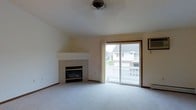 This single-floor space centers around a spacious, carpeted living area with a clean, neutral aesthetic. A corner fireplace with a tile surround and raised white mantel serves as a focal point, adding warmth and architectural interest to the room. The ceiling is smooth and slightly vaulted near the fireplace corner, lending a subtle sense of height and openness to the space. Natural light flows generously into the room through a large sliding glass door, which provides direct access to an outdoor balcony or patio area and frames views of the surrounding neighborhood. A wall-mounted air conditioning unit is positioned near the sliding door, offering climate control convenience. The neutral carpet throughout the living area complements the light-toned walls, creating a bright and cohesive environment. Overhead, a centrally mounted ceiling light fixture provides additional illumination. The overall ambiance is understated and functional, with an open layout that allows for versatile furniture arrangement. The combination of the fireplace, natural light, and outdoor access gives the space a comfortable, livable character suited to everyday use.