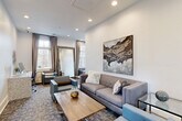 Located at 100 Cavalier Crossing in Stonecrest, Georgia, this thoughtfully designed single-floor space serves as both a comfortable lounge and a functional work area. The open layout is anchored by crisp white walls and patterned carpet flooring, creating a clean, modern aesthetic that flows seamlessly from one zone to the next.

The lounge area is furnished with a tufted gray sofa and coordinating blue accent chairs arranged around a rustic wood coffee table, offering a welcoming and well-appointed setting. A large-scale abstract canvas painting with sweeping earth tones adds a striking focal point to the main wall, while a decorative circular wall sculpture provides additional visual interest in the workstation corner.

A built-in granite-style desk accommodates dual computer workstations, complemented by adjustable task chairs and flanked by additional seating for collaborative use. A generously sized ornate floor mirror reflects light and enhances the sense of depth within the space.

Tall windows dressed with neutral draping curtains invite abundant natural light and offer views of the surrounding greenery, contributing to an airy and uplifting atmosphere. Recessed ceiling lighting ensures consistent illumination throughout. The overall ambiance is polished yet approachable, balancing professional functionality with residential comfort.