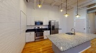 Located on University Avenue in Rochester, NY, this single-floor space showcases a compelling blend of industrial heritage and contemporary upgrades. The layout is generously proportioned, with soaring ceilings featuring exposed wooden beams and visible ductwork that lend an authentic loft-style character throughout. Warm hardwood flooring flows consistently across the main areas, complementing the crisp white walls and exposed brick accents that add texture and visual interest.

The kitchen is a standout feature, equipped with stainless steel appliances including a range, microwave, and French door refrigerator. White shaker-style cabinetry with dark countertops lines the walls, while a large granite-topped island with an undermount sink and gold-toned faucet anchors the space. Industrial-style pendant lighting hangs from the high ceiling, reinforcing the loft aesthetic.

The bedroom areas are well-proportioned with high ceilings, hardwood floors, and large windows that allow natural light to filter in. One room features a striking exposed brick accent wall. A bathroom offers tile flooring, granite vanity countertops, and accessibility-friendly grab bars. The overall ambiance is open, airy, and urban, making this a versatile space well-suited for both living and creative use.