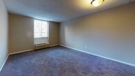Located at 402 Foulk Rd in Wilmington, Delaware, this single-floor unit offers a practical and functional layout across several well-defined rooms. The living area is generously proportioned, featuring wall-to-wall carpeting in a neutral tone, a drop-panel ceiling, and a window with a built-in HVAC unit that provides both natural light and climate control. The open flow between the main living space and adjoining rooms creates a cohesive and easy-to-navigate floor plan. The kitchen is equipped with white cabinetry extending to the ceiling for ample storage, granite-style countertops, a stainless sink, and white appliances including a range, dishwasher, and refrigerator. Dark hardwood-style flooring grounds the kitchen and adds a contrasting warmth to the space. The bedroom is well-sized with plush carpeting, neutral walls, and a standard closet with a rod and shelf for organized storage. A second carpeted room provides additional flexible space. The bathroom features wood-look flooring, a white vanity with a single sink, a mirror with overhead lighting, tiled wainscoting, and a clean, efficient layout. Throughout the unit, clean white trim and understated neutral tones contribute to a simple, move-in-ready atmosphere.