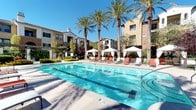 This expansive outdoor amenity area is designed for resort-style living, offering a comprehensive range of leisure features set against a backdrop of mature palm trees, manicured hedges, and lush greenery under abundant natural sunlight. The centerpiece is a generously sized swimming pool with crystal-clear blue water, flanked by rows of lounge chairs and elegant white canopy cabanas, providing both open-air sunbathing and shaded relaxation options. A separate spa with bubbling water offers a more intimate retreat, tucked beneath a pergola and surrounded by climbing greenery and vibrant potted plants.

A striking terracotta-hued outdoor fireplace with an arched glass door serves as a dramatic architectural focal point, complemented by teal-cushioned seating nearby for cozy gatherings. The outdoor kitchen area features a stainless steel built-in gas grill set into a tiled countertop beneath a pergola, with a bar-height counter and iron chairs shaded by a bold orange umbrella. The overall aesthetic reflects a warm Southwestern Mediterranean influence, with earthy tones, tile rooflines, and abundant plantings creating a cohesive, inviting atmosphere throughout.