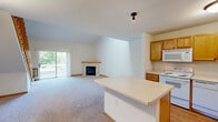 This two-floor residence offers a well-designed layout with a bright, open-concept main level. The living area is anchored by a corner fireplace with a light wood surround and carpeted flooring, while a sliding glass door provides direct access to an outdoor deck with views of surrounding greenery. A wooden staircase with a slatted railing connects the two floors and adds a warm architectural detail to the space.

The kitchen is efficiently arranged with warm oak cabinetry, white appliances including a range, dishwasher, microwave, and refrigerator, and light hardwood flooring. A peninsula island with a light countertop provides additional prep space and separates the kitchen from the living area. Open shelving near the entry adds a practical storage element.

Upstairs, the primary bedroom features plush carpeting, a ceiling fan, and direct access to a private en-suite bathroom with oak vanity cabinetry and a lighted mirror. A generously sized walk-in closet with dual hanging rods and carpeted flooring provides ample storage. An additional upper-level room features a vaulted ceiling with a skylight and ceiling fan, flooding the space with natural light. A full bathroom on this level includes a tub-shower combination, oak vanity, and tile flooring.