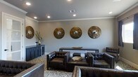 This single-floor space presents a polished and contemporary lounge area designed with both comfort and style in mind. The room is finished with smooth grey walls that serve as a neutral backdrop, accented by crisp white crown molding that adds a refined architectural detail along the ceiling line. Recessed lighting is evenly distributed throughout the ceiling, providing consistent, warm illumination across the space.

The seating arrangement centers around a collection of deep, tufted dark leather club chairs and a matching sofa, positioned to encourage conversation and relaxation. A low wooden coffee table with a round tray sits at the heart of the grouping, complementing the rich tones of the leather furniture. A sleek dark console table runs along the main wall, offering a functional surface beneath a striking display of four large, circular metallic wall medallions. These decorative pieces feature hammered bronze and silver mirror centers, serving as bold focal points that elevate the room's aesthetic.

The floor is covered in a patterned area rug with a scrolling motif in neutral tones, grounding the seating area and adding textural interest. A dark credenza along a side wall completes the room, providing additional storage. Overall, the space exudes a sophisticated, upscale ambiance.