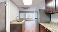 This single-floor unit offers a practical and well-appointed layout comprising a living room, kitchen, and bathroom. The kitchen is a standout feature, equipped with stainless steel appliances including a full-size refrigerator, gas range, and over-the-range microwave. Rich dark-brown cabinetry provides ample storage, complemented by granite-style countertops that add a polished finish. A stainless steel undermount sink with a modern faucet completes the functional workspace. The kitchen flows toward the living area, which features warm hardwood-style flooring and a wide window fitted with vertical blinds that allow for natural light and privacy control. The ceiling incorporates recessed panel tiles with integrated lighting, contributing to a clean, bright atmosphere throughout. The living space is finished with neutral gray walls and crisp white baseboards, creating a simple, versatile backdrop. The bathroom maintains a consistent, understated aesthetic with white square wall tiles, gray floor tiles, a wall-mounted sink, large mirror, and standard fixtures. Overall, the unit presents a functional, neatly finished space well-suited for efficient everyday living.