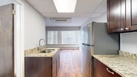 This single-floor unit offers a practical and well-appointed layout comprising a living room, kitchen, and bathroom. The kitchen is a standout feature, equipped with stainless steel appliances including a full-size refrigerator, gas range, and over-the-range microwave. Rich dark-brown cabinetry provides ample storage, complemented by granite-style countertops that add a polished finish. A stainless steel undermount sink with a modern faucet completes the functional workspace. The kitchen flows toward the living area, which features warm hardwood-style flooring and a wide window fitted with vertical blinds that allow for natural light and privacy control. The ceiling incorporates recessed panel tiles with integrated lighting, contributing to a clean, bright atmosphere throughout. The living space is finished with neutral gray walls and crisp white baseboards, creating a simple, versatile backdrop. The bathroom maintains a consistent, understated aesthetic with white square wall tiles, gray floor tiles, a wall-mounted sink, large mirror, and standard fixtures. Overall, the unit presents a functional, neatly finished space well-suited for efficient everyday living.