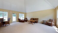 This single-story space is defined by its striking vaulted ceiling, which soars to an impressive height and lends a sense of grandeur and airiness to the living area. The ceiling is finished in crisp white, contrasting gently with the warm, pale yellow walls that create a calm and welcoming atmosphere throughout. Soft, neutral carpeting covers the floor, adding comfort underfoot and contributing to the room's understated, relaxed aesthetic.

Large windows along one wall, complemented by a glass-paneled exterior door, allow generous amounts of natural light to flood the interior while offering views of the lush greenery outside. Rattan and wicker furnishings, including a sofa, armchairs, and a wooden coffee table, are arranged to create a comfortable and casual seating area suited to relaxed gatherings.

The overall ambiance is light, open, and uncluttered, with the soaring roofline serving as the architectural centerpiece of the space. The combination of high ceilings, natural light, and simple, warm finishes gives the room a breezy, informal character that feels both spacious and inviting.