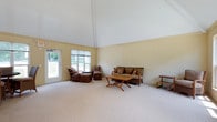 This single-story space is defined by its striking vaulted ceiling, which soars to an impressive height and lends a sense of grandeur and airiness to the living area. The ceiling is finished in crisp white, contrasting gently with the warm, pale yellow walls that create a calm and welcoming atmosphere throughout. Soft, neutral carpeting covers the floor, adding comfort underfoot and contributing to the room's understated, relaxed aesthetic.

Large windows along one wall, complemented by a glass-paneled exterior door, allow generous amounts of natural light to flood the interior while offering views of the lush greenery outside. Rattan and wicker furnishings, including a sofa, armchairs, and a wooden coffee table, are arranged to create a comfortable and casual seating area suited to relaxed gatherings.

The overall ambiance is light, open, and uncluttered, with the soaring roofline serving as the architectural centerpiece of the space. The combination of high ceilings, natural light, and simple, warm finishes gives the room a breezy, informal character that feels both spacious and inviting.
