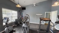 This single-story community clubhouse is designed to offer residents a welcoming and functional gathering space. The grand lobby greets visitors with soaring ceilings, recessed lighting, and a sophisticated chandelier that casts a warm glow over the open layout. Dark-trimmed double doors with glass panes flood the entrance with natural light, complemented by neutral tones and tasteful décor including fresh florals and curated artwork.

The lounge area serves as a standout feature, centered around a floor-to-ceiling stacked stone fireplace with a rustic wood mantel. Comfortable seating, including upholstered sofas and wingback chairs, creates an inviting atmosphere for relaxation or casual conversation. French doors open to an outdoor area, enhancing the sense of connectivity between interior and exterior spaces.

A secondary seating area continues the refined aesthetic, featuring cream-toned sofas accented with soft pillows, elegant chandeliers, and glass-paneled French doors that allow abundant natural light to filter through.

The fitness center is well-equipped for residents seeking an active lifestyle, housing elliptical machines, a treadmill, a dumbbell rack, and medicine balls. A wall-mounted television, pendant lighting, a ceiling fan, and window blinds complete this practical and comfortable workout space.