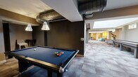 This single-floor amenity space is thoughtfully designed to accommodate both relaxation and entertainment across a generous footprint. The layout flows seamlessly between two distinct zones, each with its own character and purpose.

The living room area exudes a modern, welcoming ambiance, featuring patterned carpet tile flooring in neutral grey tones throughout. Comfortable seating arrangements include a tufted grey sofa, vibrant teal accent chairs, and a sleek white coffee table, all set beneath warm recessed and pendant lighting. A dining nook with a round table and mid-century style chairs adds versatility, while curated wall art and decorative gold wall accents contribute to the polished aesthetic. The space benefits from high ceilings and natural light filtering in through clerestory windows.

The game room offers an energetic counterpoint, anchored by a full-size pool table with a blue felt surface and pendant lighting overhead. Exposed industrial-style ductwork on the ceiling adds an urban edge to the darker, moodier atmosphere. A shuffleboard table, wall-mounted flat-screen televisions, a cue rack, and a bar counter with stools complete the entertainment-focused amenities. Together, these two spaces create a dynamic and inviting environment suited for both casual gatherings and active recreation.