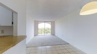 This single-floor space offers a clean, minimalist layout filled with abundant natural light. The main living area is defined by soft, neutral-toned carpeting that transitions seamlessly into light ceramic tile flooring near the entryway, creating a clear and functional division of space. The walls are finished in a crisp white throughout, contributing to an open and airy ambiance that feels both welcoming and versatile.

A standout feature of the living area is the large sliding glass door framed by an elegant arched window above, allowing generous amounts of natural light to pour into the room. This architectural detail adds a touch of character to an otherwise understated design. The sliding door opens directly onto a private balcony enclosed by a classic wrought-iron railing, offering picturesque views of lush, mature greenery beyond — a serene backdrop that brings the outdoors in.

The ceiling is smooth and well-proportioned, enhancing the sense of spaciousness throughout. A partial countertop or ledge is visible at the edge of the space, hinting at an adjacent kitchen or dining area. Overall, the space presents a bright, neutral, and move-in-ready environment well-suited for comfortable everyday living.