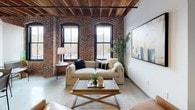 This 2-bedroom, 1-bathroom loft-style residence masterfully combines historic industrial character with contemporary living. Exposed red brick walls and rich wooden beam ceilings run throughout, creating a warm and textured ambiance that defines the space's identity.

The living area is anchored by large arched windows with black frames that flood the room with natural light and offer views of the surrounding urban streetscape. The dining area continues the industrial aesthetic, with exposed brick accents complementing a sleek marble-top table beneath warm overhead lighting.

The kitchen is efficiently designed, outfitted with stainless steel appliances, gray shaker cabinetry, and dark stone countertops — a clean, functional space that balances style with practicality.

Both bedrooms carry the hallmark exposed brick and wood-beam ceilings, each featuring arched windows that invite natural light and urban views. Dedicated closets provide ample storage in each room, and the warm palette creates a cozy, inviting atmosphere.

The bathroom maintains the building's signature character with exposed brick walls, black-framed windows, a gray vanity with stone countertops, and a warm wood-framed mirror, tying together the industrial-modern aesthetic found throughout the home.