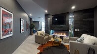 Located at 9 Tower Lane in New Haven, CT, this single-floor amenity lounge is a masterclass in contemporary interior design. The space is defined by a bold, dark color palette—deep charcoal walls, dark tile accents, and light hardwood flooring—creating a dramatic yet inviting atmosphere throughout.

At the heart of the lounge sits a striking floor-to-ceiling fireplace clad in dark metallic tile, topped with a wall-mounted television, and flanked by comfortable lounge seating arranged over cowhide rugs. A second, more expansive seating area features a large wraparound sectional sofa in deep teal velvet, accented by a sculptural sputnik chandelier and complementary gold coffee tables, adding warmth and personality to the space.

A dedicated game area includes a round poker-style table with leather seating, positioned near a large industrial-style glass garage door that opens to the outdoors, flooding the area with natural light. Shimmering chain curtains serve as elegant room dividers, subtly separating functional zones while maintaining an open, flowing layout.

The built-in wet bar and kitchen area features dark cabinetry, sleek countertops, and a vibrant teal hexagonal tile backsplash, complete with an under-counter oven and wall-mounted display. Curated artwork and lush indoor plants add the finishing touches to this thoughtfully designed communal space.