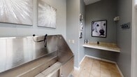 Located at 215 Rocky Ridge Blvd in Douglasville, Georgia, this single-floor space is purpose-built as a dedicated pet grooming and washing facility. The room is finished with cool gray walls and neutral tile flooring, creating a clean, functional environment with an inviting aesthetic.

The centerpiece of the space is a large, professional-grade stainless steel dog bathing station equipped with a handheld sprayer and leash restraint attachments, designed for ease of use and thorough pet care. Adjacent to the wash station is a built-in granite-topped counter, ideal for grooming tools and supplies, accented by dog-themed artwork and décor that adds personality to the space.

A set of tall French doors lines one wall, flooding the room with abundant natural light and offering views of the surrounding greenery and parking area outside. The doors also provide convenient direct access to the exterior. The elevated ceiling height enhances the sense of openness within the compact footprint.

Wall-mounted artwork featuring canine photography and abstract prints reinforces the pet-focused theme throughout. The combination of practical fixtures, thoughtful finishes, and welcoming décor makes this a well-appointed space suited for professional or community pet grooming use.