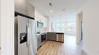 This thoughtfully designed single-floor residence in Duluth, Georgia, offers a contemporary living experience with a clean, modern aesthetic throughout. The open-concept kitchen is a standout feature, showcasing two-tone cabinetry with white upper cabinets and gray lower cabinets accented by gold hardware, a classic white subway tile backsplash, and a full suite of stainless steel appliances including a French door refrigerator, electric range, over-the-range microwave, and dishwasher. Pendant lighting hangs above the kitchen peninsula, which flows seamlessly into the bright living area, where large windows and a ceiling fan create an airy, welcoming atmosphere. Light wood-look flooring runs throughout the main living spaces, adding warmth and continuity. The bedrooms are finished with plush carpeting and feature generous natural light from double windows with blinds. The primary suite includes a spacious walk-in closet outfitted with wire shelving, and an en-suite bathroom with a dual-sink vanity, slate-blue cabinetry, and wood-look flooring. Five-panel gray interior doors provide a consistent, modern design element throughout. A private balcony with a dark metal railing offers views of the surrounding greenery and neighborhood streetscape.