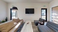 This single-floor space is designed with an open-concept layout that integrates the kitchen, dining, and living areas into one cohesive environment. The kitchen is well-appointed with stainless steel appliances, including a full-size refrigerator with an ice maker, a gas range with an overhead vent hood, and a microwave. White shaker-style cabinetry with brushed metal hardware lines the walls, complemented by gray countertops and a double-basin sink positioned beneath a window that allows natural light to enter the space. A peninsula with bar-height seating extends from the counter, providing an additional casual dining option alongside a dedicated round dining table with chairs. Wood-look flooring runs throughout, contributing to a warm and cohesive aesthetic. The living area offers multiple seating arrangements, including a leather sofa, gray upholstered chairs, and a slab-style coffee table, all oriented around a wall-mounted flat-screen TV. Windows on multiple walls bring in natural light and views of the surrounding exterior. Exit signage and the overall layout suggest this space is designed for communal or shared use, with a clean, modern, and functional design throughout.