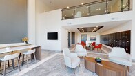 Located at 140 E Monument Ave in Dayton, Ohio, this contemporary lobby presents a sophisticated first impression through its spacious, open layout and refined modern design. The space is anchored by dramatic double-height ceilings that rise to an upper-level mezzanine, enclosed by sleek glass-panel railings, adding a sense of vertical grandeur to the interior.

The flooring throughout is a polished marble-look tile laid in a large-format pattern with subtle inlay detailing, lending an upscale, cohesive aesthetic across the entire floor. Large, floor-to-ceiling windows at the entrance flood the space with natural light while offering views of the surrounding streetscape.

Multiple distinct seating zones define the lobby's layout. A central lounge area features neutral white accent chairs arranged around organic-shaped wooden coffee tables, creating a relaxed gathering space. A secondary seating cluster showcases bold red chairs paired with a circular side table, adding a vibrant contrast against the light palette. A built-in bar-height work surface with white counter stools and a wall-mounted television provides a functional workspace area.

A wall of resident mailboxes lines one side of the lobby, while curated artwork, recessed tray ceiling lighting, and carefully placed greenery contribute to the polished, welcoming ambiance of the space.