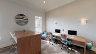 Located at 20155 San Joaquin Terr in Ashburn, Virginia, this single-floor business center offers a thoughtfully designed workspace that balances functionality with contemporary style. The space features soaring ceilings accented by elegant crown molding and is finished with soft, neutral-toned walls that create a bright and professional atmosphere.

The main workspace area includes a long, natural wood workstation desk outfitted with multiple computer monitors and ergonomic task chairs, providing an efficient environment for productivity. Decorative wall sconces offer warm ambient lighting, complemented by natural light streaming through large windows fitted with white blinds. A geometric-patterned carpet in muted gray tones anchors the space and adds visual interest underfoot.

A comfortable teal lounge chair with an arc floor lamp provides a relaxed seating option alongside the workstations. A small bistro table with two white chairs offers an additional informal meeting or break area, set beneath a striking teal world map canvas that serves as a focal point on the wall.

A wall-mounted flat-screen television enhances the space's versatility for presentations or media viewing. A central wood-toned counter/bar-height table near the entry area, adorned with a bold circular abstract artwork, rounds out the welcoming and multi-functional design. The overall ambiance is polished, modern, and well-suited for both focused work and casual collaboration.