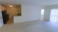 This single-floor space offers a clean, functional layout designed for comfortable everyday living. The main living area is expansive and open, finished with plush wall-to-wall carpeting that adds warmth and softness underfoot. Crisp white walls throughout create a bright, neutral backdrop that makes the space feel airy and inviting.

A pass-through kitchen counter connects the kitchen to the living area, promoting an open-concept feel while maintaining a defined cooking space. The kitchen is equipped with white cabinetry and features a sink positioned along the counter, providing practical workspace. The raised breakfast bar-style counter offers a casual dining or workspace option.

Natural light filters into the space through a window visible toward the far end of the unit, brightening the interior and drawing the eye toward what appears to be an adjacent room or hallway beyond. The overall layout flows smoothly from the kitchen into the living area and onward, making the space feel connected and cohesive.

The unit's straightforward design, neutral color palette, and carpeted flooring create a versatile canvas well-suited for a variety of furnishing styles. The combination of open living space, functional kitchen access, and natural light makes this a practical and comfortable single-floor residence.