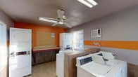 This single-story laundry facility is designed to provide a practical and functional space for shared use. The interior is distinguished by a bold two-tone color scheme, featuring vibrant orange accent walls paired with gray upper walls and a horizontal orange stripe running along the mid-wall, creating a visually cohesive environment. The lower portion of the walls is finished with neutral tile wainscoting, complementing the durable tile flooring throughout.

The facility is equipped with multiple top-loading washing machines and a stacked washer-dryer unit, offering ample capacity for residents. A dedicated folding counter runs along one wall, providing a convenient surface for sorting and folding laundry. A bulletin board is mounted nearby for informational postings and service instructions.

Overhead fluorescent lighting ensures the space is well-lit, while a ceiling fan promotes air circulation. A wall-mounted air conditioning unit provides additional climate control for year-round comfort. Natural light enters through a window fitted with blinds, adding brightness to the space.

Access to the facility is through a keyed entry door, ensuring security for users. Overall, the space is straightforward and well-equipped, prioritizing function and ease of use.