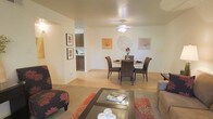 This single-floor residence showcases an open-concept layout that seamlessly integrates the living, dining, and kitchen areas into one cohesive space. The living area features warm carpeting and is furnished with comfortable seating, including a floral-patterned accent chair and a sleek glass-top coffee table set atop a decorative area rug. Transitioning into the dining area, light wood laminate flooring creates a clean, distinct boundary between the two zones. A round dining table surrounded by woven rattan-style chairs serves as the centerpiece of this space, topped with a simple floral arrangement that adds a natural accent. A ceiling fan with integrated lighting is centrally positioned overhead, providing both ventilation and ambient illumination. The walls are adorned with framed artwork and botanical-inspired wall décor, contributing a warm, welcoming atmosphere throughout. A hallway opening in the background leads to the kitchen, where dark wood cabinetry is visible, hinting at additional storage and functional workspace beyond. The overall ambiance is bright, casual, and comfortably livable.