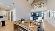 Located at 5532 38th St S in Fargo, North Dakota, this single-floor clubhouse is a thoughtfully designed community gathering space that balances modern aesthetics with warm, inviting materials. The defining architectural feature is the dramatic vaulted ceiling accented by exposed natural wood trusses, which lend a rustic-contemporary character to the open interior. Herringbone-patterned hardwood flooring flows throughout, adding visual texture and a refined finish underfoot.

The main lounge area is anchored by a substantial reclaimed wood console table and complemented by a dark leather sofa with plaid accent pillows, creating a relaxed yet polished seating arrangement. A teal feature wall serves as a striking backdrop for a wall-mounted television above a sleek electric fireplace, flanked by modern white lounge chairs. Floor-to-ceiling black-framed glass panels at the entrance flood the space with natural light while offering a clean, industrial-chic design detail.

A staffed reception desk with warm wood cabinetry provides a welcoming point of service, while a dedicated refreshment station features white shaker cabinetry, dark countertops, gold hardware, subway tile, and a coffee machine — ideal for resident convenience. A hallway connects to additional rooms, completing this well-appointed and functional clubhouse.