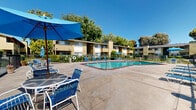 Located at 6184 Cottle Road in San Jose, California, this outdoor amenity area centers around a generously sized swimming pool with clear, blue water and a distinctive brick-edged surround. The pool area is designed for both relaxation and recreation, featuring multiple entry points with stainless steel handrails for easy access.

The expansive concrete sun deck surrounding the pool is well-appointed with numerous blue lounge chairs and chaise lounges, offering ample space for sunbathing. Several patio table-and-chair sets with blue shade umbrellas are positioned throughout the deck, providing comfortable spots for outdoor dining or socializing in the shade.

To one side of the pool, a covered clubhouse or pavilion structure offers a shaded gathering space, extending the usability of the outdoor area throughout the day. A safety life ring is also visibly stationed near the pool perimeter.

The entire pool area is enclosed by a black iron fence, providing both security and a clear boundary. The surrounding landscape features mature trees and well-maintained flowering shrubs that frame the space with natural greenery. The multi-story residential buildings surrounding the courtyard are finished in warm earth tones, contributing to a cohesive and inviting community aesthetic. Under the clear California sky, this outdoor space offers a bright, open, and welcoming environment.