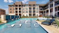 Located at 1160 Montgomery Dr in Allen, TX, this outdoor amenity space is designed to deliver a resort-style experience for its residents. The centerpiece is a generously sized swimming pool featuring sparkling blue water, decorative water wall features with cascading tile accents, and a shallow wading area furnished with sleek, modern white in-pool lounge chairs — ideal for relaxing under the Texas sun. The pool deck is thoughtfully designed with a combination of concrete, artificial turf, and stone-accented pavers, creating distinct zones for lounging, socializing, and recreation. Surrounding the pool, residents will find multiple seating arrangements, including classic chaise lounges and round patio tables with chairs shaded by striped umbrellas. Adjacent to the pool area is a covered outdoor pavilion supported by natural limestone columns and topped with a standing-seam metal roof. This structure houses a built-in stainless steel grill, outdoor kitchen counter space, and a large mounted flat-screen television, making it a prime gathering spot for outdoor entertaining. Lush landscaping with native plants and mature trees frames the perimeter, providing privacy and a natural aesthetic. The multi-story residential building overlooks the amenity area, offering residents on upper-floor balconies elevated views of this vibrant outdoor space.
