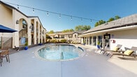 Located at 10038 Regal Park Ln in Dallas, TX 75230, this generously sized single-level outdoor amenity complex offers a resort-like experience centered around a large, sparkling swimming pool. The pool area is framed by a wide, smooth concrete deck that provides ample space for sunbathing and socializing, furnished with wicker lounge chairs accented by yellow cushions and shaded dining sets topped with navy blue umbrellas. String lights draped overhead lend a welcoming ambiance for both daytime and evening use. A dedicated outdoor lounge zone features comfortable sectional seating surrounded by lush tropical and ornamental plantings, offering a quiet retreat from the pool deck. The adjacent single-story clubhouse building features a sloped shingle roof, large glass panel windows, and multiple entry doors that invite natural light deep into the interior, which appears to include a fitness area visible from the pool side. The surrounding multi-story residential buildings showcase a stucco Mediterranean-influenced architectural style with arched entryways, wrought iron railings, and classic shuttered windows. The entire space is enclosed by decorative iron fencing and accented by mature trees and vibrant greenery, creating a private, resort-caliber outdoor environment. Two bathrooms serve the space.