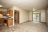 This single-floor residential unit offers a practical and comfortable layout designed for everyday living. The open-concept living and dining area is anchored by plush carpeting and transitions seamlessly into a tiled entryway and kitchen zone. A ceiling fan overhead and a sliding glass door leading to a private balcony fill the living space with natural light and provide pleasant outdoor views of the surrounding greenery. The kitchen is well-appointed with warm oak cabinetry, dark laminate countertops, a double stainless steel sink with under-cabinet lighting, and a freestanding electric range, offering ample storage and prep space. A secondary living or flex area connects to the kitchen, providing additional versatile space. The two bedrooms are carpeted and feature neutral walls with wood-trimmed windows that let in natural light; one bedroom includes a walk-in closet with built-in shelving for added storage convenience, while the other features a built-in window bench. The full bathroom is finished with tile flooring, a wood-trimmed vanity with a light stone-tone countertop, a large mirror, and a glass-door shower and tub combination. Throughout the unit, warm wood door and window trim provides a cohesive, classic aesthetic.