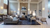 This single-floor communal space is defined by its grand architectural presence and carefully curated interior design. The main lounge area commands attention with dramatically tall ceilings framed by white coffered detailing and bold structural columns. A floor-to-ceiling stacked stone accent wall anchors the room, centered around a classic fireplace with a white mantel, creating a refined yet welcoming focal point. Light-toned herringbone-patterned flooring flows continuously throughout, lending visual cohesion and an air of sophistication to the entire space.

The lounge is furnished with generous sectional sofas, accent chairs in navy and neutral tones, and layered area rugs that define distinct seating arrangements. Contemporary abstract artwork adorns the walls, complementing the cool gray and blue color palette. A dedicated dining area features a long communal table surrounded by wood-and-cane chairs, positioned near tall multi-pane windows that invite abundant natural light and views of surrounding greenery.

A separate lounge nook offers a more intimate setting, featuring a dramatic dark textured stone feature wall, wall-mounted television, and compact seating with a small café-style counter — ideal for casual gatherings. Warm sconce lighting throughout enhances the inviting ambiance of this well-appointed space.