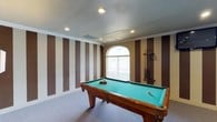 Located at 147 Links Dr in Canton, Mississippi, this single-floor space makes a memorable impression with its thoughtfully designed interior. The highlight of the space is a dedicated game room that combines function and style in equal measure. Bold vertical stripes in alternating tan and chocolate brown command the walls, creating a dynamic and energetic atmosphere that sets the tone for recreation and entertainment. A full-size pool table with a vibrant teal felt surface anchors the room as its centerpiece, accompanied by a wall-mounted cue rack keeping equipment neatly organized and within reach. An arched window allows natural light to filter into the room, softening the bold décor and providing a touch of architectural elegance. Recessed lighting in the textured ceiling ensures the space is well-lit for evening use. Soft carpeting underfoot adds comfort and warmth to the room's overall feel. Crown molding along the ceiling line adds a refined finishing touch, bridging the playful design elements with a sense of classic detail. Altogether, this space offers an inviting and well-appointed environment ideal for leisure and entertaining.
