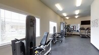 This fitness center is designed to support a comprehensive workout experience within a single-floor layout. The space features vaulted ceilings that lend an open, airy feel, while multiple windows fitted with horizontal blinds allow generous natural light to fill the room, creating a bright and motivating environment.

The flooring throughout is durable rubber, a practical choice that provides both cushioning and stability for exercise activities. A selection of strength training machines is arranged along the room, including leg press and cable resistance equipment, offering users a range of targeted muscle group exercises. Additional multi-station cable machines are positioned toward the rear of the space, maximizing the available floor area.

Overhead fluorescent lighting supplements the natural light, ensuring the space remains well-lit at all times of day. A mirrored wall section at the far end of the room reflects the equipment and expands the perceived depth of the space, while also serving a functional role for form-checking during workouts.

The neutral, warm-toned walls and clean layout contribute to a focused and professional atmosphere. The overall design prioritizes functionality and ease of movement, making this fitness center a practical and well-appointed facility for strength and conditioning training.