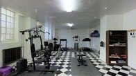 This dedicated fitness space is designed to support a comprehensive workout experience within a single-floor layout. The room features a striking black-and-white checkered tile floor, giving it a bold, classic aesthetic that stands out against the clean white walls. Overhead, flush-mounted lighting and a ceiling fan work together to keep the space well-lit and well-ventilated during workouts.

The room is outfitted with a diverse selection of exercise equipment, including a multi-station strength training machine, a stationary cycle, two treadmills, and an elliptical trainer, providing ample options for both cardio and resistance training. A dedicated free-weight and bench area is also present, complemented by a wall-mounted rack stocked with a variety of medicine balls and weighted exercise balls, catering to functional and core training routines. Rubber floor mats are strategically placed beneath key equipment to provide stability and protect the flooring.

Natural light filters in through a window on one side of the room, adding brightness to the space. The overall layout is practical and spacious, with equipment arranged to allow comfortable movement between stations. This fitness room offers a well-rounded, functional environment suited for a full range of exercise activities.