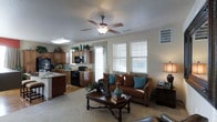 This single-floor space showcases an open-concept layout that connects the living area and kitchen, creating a cohesive and functional environment. The living room is anchored by a warm brown sofa accented with teal throw pillows, complemented by a classic dark-wood coffee table that adds a touch of traditional elegance. A ceiling fan with a built-in light fixture is centrally positioned overhead, enhancing air circulation and providing ambient lighting. Multiple windows fitted with white blinds allow generous natural light to fill the room, contributing to its bright and welcoming atmosphere. The kitchen flows naturally from the living space and features warm-toned wood cabinetry, a black range, a built-in microwave, and a kitchen island with a sink, offering both style and practicality. A skylight above the kitchen area further brightens the workspace. The flooring transitions from carpet in the living area to a hard surface in the kitchen, clearly delineating the two functional zones. Together, the warm color palette, thoughtful layout, and ample natural light create a comfortable and inviting home environment ideal for both everyday living and entertaining.