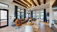 Located at 13555 Yellow Fern Way in Riverview, Hillsborough County, Florida, this single-floor community clubhouse delivers a sophisticated and well-appointed gathering space. The main lounge is anchored by soaring ceilings with prominent dark wood beam accents and a striking multi-bulb brass chandelier. Large black-framed windows and glass doors flood the space with natural light while offering outdoor views, and teal drape panels add a bold color contrast against the crisp white walls. A dramatic floor-to-ceiling brick fireplace serves as a central focal point, surrounded by elegantly arranged seating including navy accent chairs and a circular wood coffee table. Light wood-look tile flooring flows consistently throughout. A secondary lounge area features a tray ceiling with a warm wood finish, deep teal accent walls, curated artwork, and inviting seating arrangements ideal for relaxed conversation. The kitchenette and entertainment zone is outfitted with dark cabinetry, a teal glass-tile backsplash, a wall-mounted TV, stainless steel appliances, pendant lighting, and a large center island with bar seating. A wooden shuffleboard table adds a recreational element nearby. Together, these spaces create a cohesive, design-forward environment well-suited for community gatherings and social events.