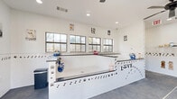 Located at 195 E Round Grove Rd in Lewisville, Texas, this single-floor pet care facility is thoughtfully designed to accommodate both professional grooming and self-serve dog washing. The space features a bright, white interior with clean subway tile wainscoting adorned with playful paw print and bone motifs, creating a welcoming atmosphere for pets and their owners alike. Whimsical dog-themed wall art, including painted breed portraits and vintage-style signs, adds personality throughout.

The bathing area is the centerpiece of the facility, showcasing raised, step-accessible wash stations with dark countertops, retractable spray hoses, and easy-clean surfaces. Multiple windows with black frames line the walls, flooding the space with natural light and providing outdoor views. The grooming area is equipped with adjustable professional grooming tables and wall-mounted dryer arms, along with a dedicated mini-split HVAC unit for climate control. A ceiling fan overhead ensures comfortable airflow across the open floor plan.

The durable concrete flooring is practical and easy to maintain, while an entry door with a glass panel adds to the brightness of the space. A seating bench, storage shelving, and waste disposal stations are thoughtfully integrated throughout, making this a fully functional and welcoming environment for pet care services.