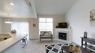 This two-story residence makes a strong first impression with its soaring vaulted ceilings in the main living area, which lend the space a sense of openness and grandeur. The living room serves as the focal point of the home, anchored by a clean, tile-surround corner fireplace that adds warmth and a classic architectural detail. A large, multi-pane window positioned high on the wall floods the room with natural light while framing outdoor views, complementing the neutral palette of white walls and light-toned carpet. The open layout allows the living area to flow naturally into an adjacent dining space, visible to the side, creating a cohesive and functional floor plan ideal for both daily living and entertaining. The two-story design adds vertical dimension and visual interest throughout, while the neutral tones and clean lines give the home a timeless, versatile aesthetic. The combination of carpeted living areas and tiled surfaces near the fireplace provides both comfort and practicality. Overall, the space offers a bright, welcoming atmosphere with well-considered architectural features.