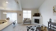 This two-story residence makes a strong first impression with its soaring vaulted ceilings in the main living area, which lend the space a sense of openness and grandeur. The living room serves as the focal point of the home, anchored by a clean, tile-surround corner fireplace that adds warmth and a classic architectural detail. A large, multi-pane window positioned high on the wall floods the room with natural light while framing outdoor views, complementing the neutral palette of white walls and light-toned carpet. The open layout allows the living area to flow naturally into an adjacent dining space, visible to the side, creating a cohesive and functional floor plan ideal for both daily living and entertaining. The two-story design adds vertical dimension and visual interest throughout, while the neutral tones and clean lines give the home a timeless, versatile aesthetic. The combination of carpeted living areas and tiled surfaces near the fireplace provides both comfort and practicality. Overall, the space offers a bright, welcoming atmosphere with well-considered architectural features.