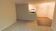 This single-floor residence offers a functional and well-organized open-concept layout designed to maximize the sense of space and ease of movement. The living area features neutral-toned wall-to-wall carpeting that creates a warm, comfortable atmosphere, transitioning seamlessly into hardwood-style flooring in the adjoining area, adding visual contrast and definition between spaces. The kitchen is thoughtfully positioned behind a half-wall pass-through counter, which serves as both a practical workspace and a visual divider while maintaining an open connection to the living space. The kitchen is equipped with white cabinetry offering ample storage, dark countertops, and a range with an overhead ventilation hood. The cabinetry layout is clean and efficient, making good use of the available space. Warm, ambient lighting throughout the unit contributes to an inviting atmosphere, while white baseboards provide a crisp finish along the walls. The neutral color palette of beige and warm gray tones throughout the space lends itself well to a variety of interior design styles, making it highly adaptable for any occupant. Overall, the layout is practical and cohesive, ideal for everyday living.